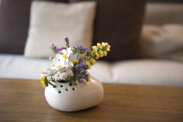 La Petra, un pequeño jarrón de cerámica blanca lleno de una variedad de flores de colores, se encuentra sobre una mesa de madera contra un fondo borroso de cojines beige y marrón.