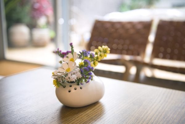 Un pequeño jarrón blanco llamado "Petra" lleno de una mezcla de flores de colores se encuentra sobre una mesa de madera, enmarcado por el fondo borroso de dos sillas tejidas y una vista exterior a través de una ventana.