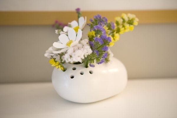 Petra, un pequeño jarrón de cerámica blanca con agujeros en la parte superior, sostiene un arreglo de flores blancas, amarillas y violetas sobre una superficie de color claro.