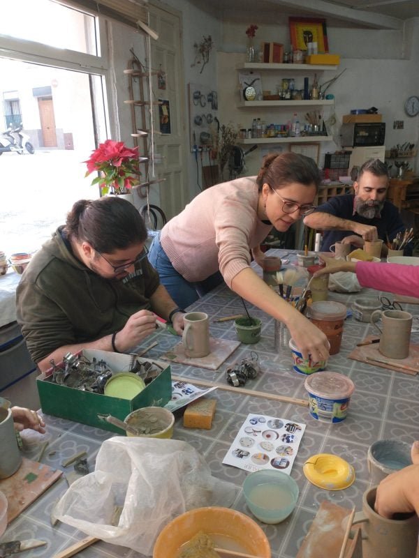 La gente pinta cerámica en una mesa de un taller con materiales y herramientas de arte esparcidos por todas partes. Una mujer está ayudando a un grupo de participantes enfocados. Al fondo se ven estanterías con diversos objetos.