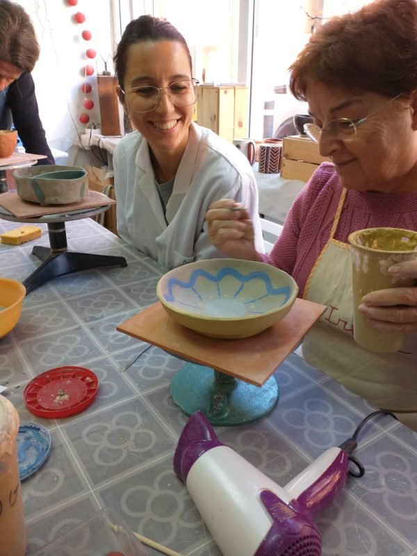 Dos mujeres en una clase de cerámica; uno pinta un cuenco, y el otro, con gafas y bata blanca, sonríe mientras observa. Sobre la mesa se ven varios objetos de cerámica y herramientas.