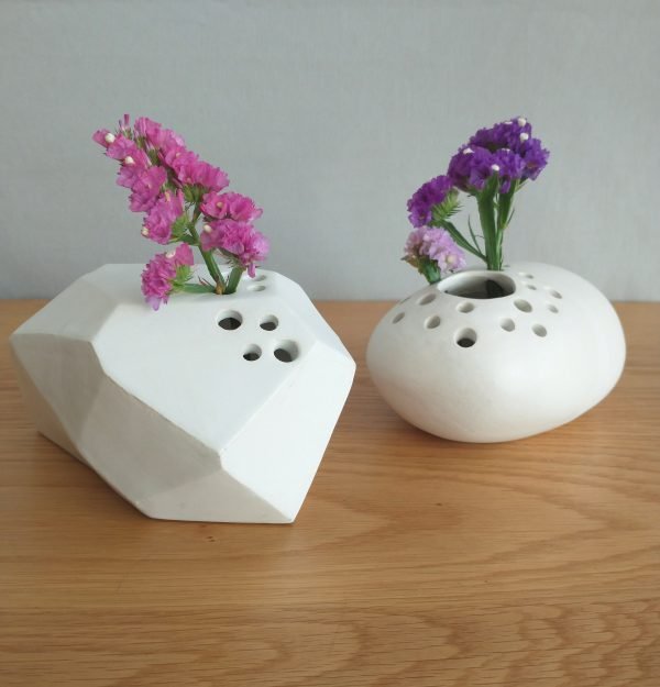 Dos jarrones Petra de cerámica blanca con formas geométricas sostienen pequeñas flores rosas y violetas sobre una superficie de madera.